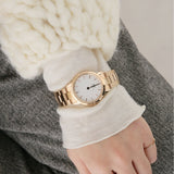 Gold wristwatch on a wrist with white sweater and grey pants.