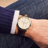 Man's wrist with a brown leather watch, wearing a blue sweater and jeans.